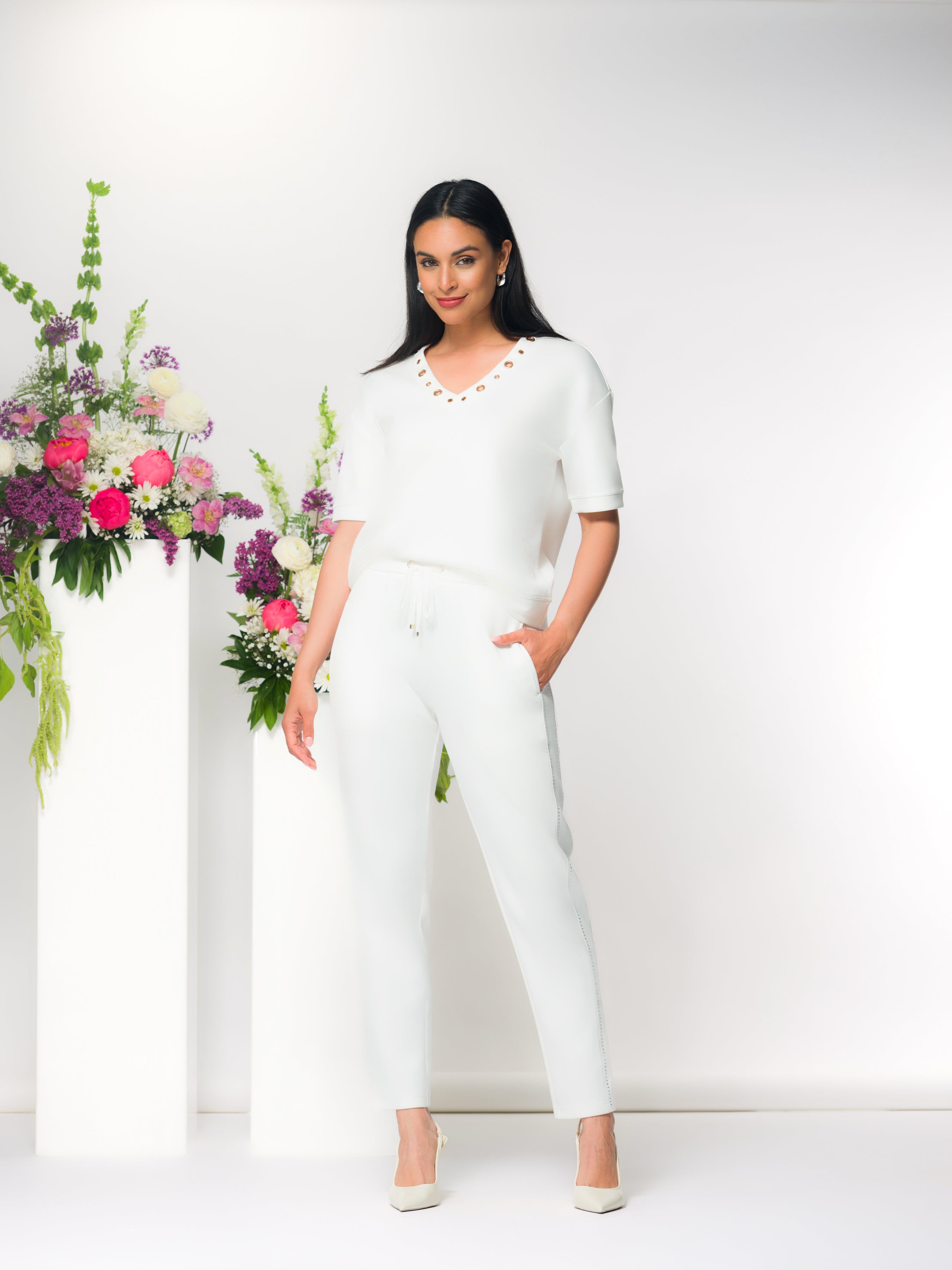 Woman wearing a relaxed white V-neck top with short sleeves and matching wide-leg pants, styled indoors with floral arrangements.