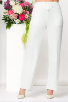 Woman with dark hair wearing white wide-leg lounge pants with a drawstring waist, standing in a minimalist studio with floral arrangements behind her