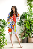 Model wearing a relaxed women’s blouse with a button-front design and short sleeves, styled with coordinating bottoms and flat sandals, standing indoors among plants.