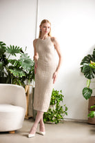 Model wearing a sleeveless textured knit midi dress with a streamlined fit, styled with heels, standing in a bright interior space with indoor plants and modern furnishings.