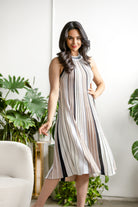 Model wearing the Hartridge Pleated Knit Dress in a neutral striped pattern featuring beige, cream, and black tones, styled with a sleeveless high neckline and flowing midi-length pleated skirt indoors.