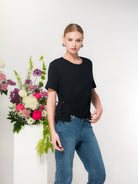 Model wearing a black short-sleeve crew neck t-shirt with subtle side detail, paired with blue jeans, standing beside a floral arrangement.