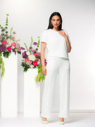 Model wearing a white short-sleeve asymmetric blouse with wide-leg white trousers and ivory heels, styled against a neutral studio background with floral arrangements.