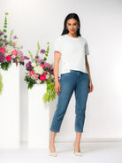 Model wearing a white short-sleeve asymmetric blouse with cropped blue straight-leg jeans and ivory pointed heels, styled in a bright studio with pink and white flowers.