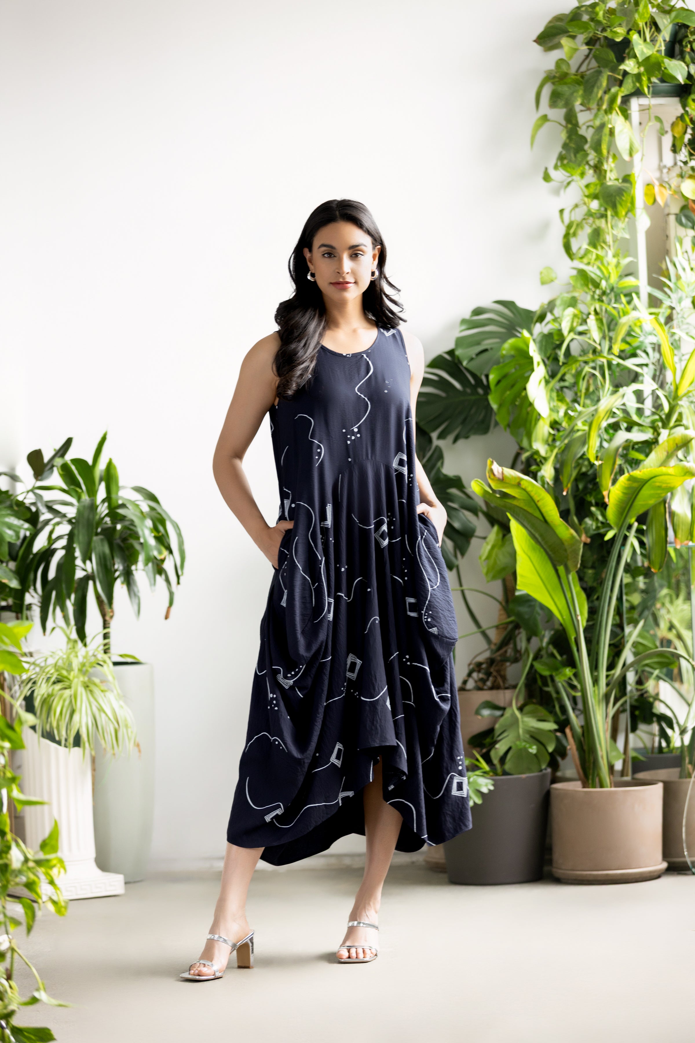 Model wearing a navy sleeveless midi dress with abstract white print, relaxed draped silhouette and side pockets, styled with silver heeled sandals, standing in a bright indoor space with green plants.