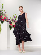 Model wearing a navy sleeveless midi dress with abstract white print and flowing asymmetrical hem, paired with cream pointed heels, photographed beside a white pedestal with colourful floral arrangement.