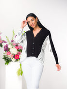 Woman modelling a mixed-media button-front sweater blouse with a V-neckline and relaxed silhouette. The design features a soft knit front panel paired with a lightweight woven abstract print panel and sleeves. Styled with slim trousers, she stands beside a floral arrangement in a bright studio setting.