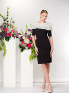 Model wearing a black knee-length sheath dress with a sheer white ruffle neckline and elbow-length sleeves, styled with nude pointed heels, standing beside tall white pedestals topped with pink and white floral arrangements.