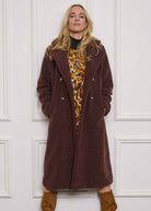 Woman wearing a long brown coat over a leopard print top against a white paneled wall.