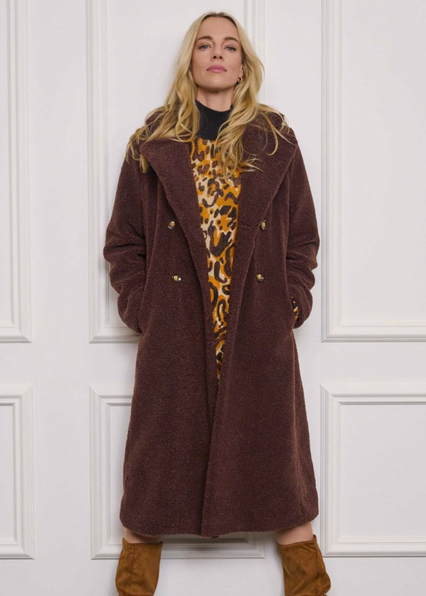 Woman wearing a long brown coat over a leopard print top against a white paneled wall.