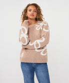 Close-up of a woman wearing a sand patterned crew-neck sweater, highlighting the knit texture, abstract motif, ribbed neckline, and cuffs.