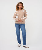Woman wearing a sand patterned crew-neck sweater with straight-leg jeans and casual sneakers, standing against a light studio background.