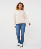 Woman wearing a beige patterned crew-neck sweater with straight-leg jeans and casual sneakers, standing against a light studio background.