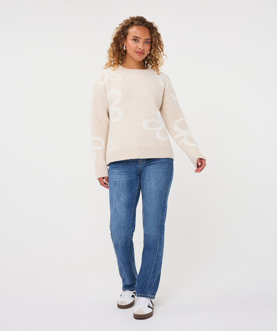 Woman wearing a beige patterned crew-neck sweater with straight-leg jeans and casual sneakers, standing against a light studio background.