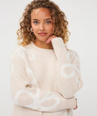 Close-up of a woman wearing a beige patterned crew-neck sweater, highlighting the knit texture, abstract motif, ribbed neckline, and cuffs.
