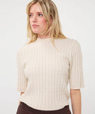 Woman modelling an ivory ribbed knit short-sleeve top with a high neckline and fitted silhouette, shown from the front.