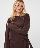 Woman modelling the Sloane Side-Tie Sweater in chocolate brown, featuring a relaxed fit, long sleeves, crew neckline, and side tie detail at the waist.