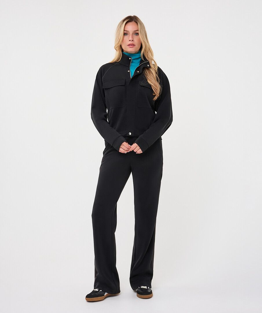 Woman wearing a black utility jacket with a stand collar and flap chest pockets over a turquoise knit top, paired with matching trousers.