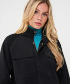 Woman wearing a black utility jacket with a stand collar and flap chest pockets over a turquoise knit top