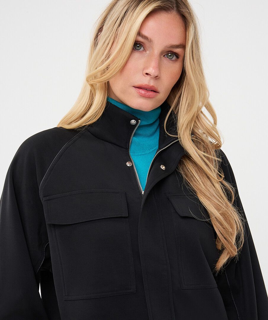 Woman wearing a black utility jacket with a stand collar and flap chest pockets over a turquoise knit top