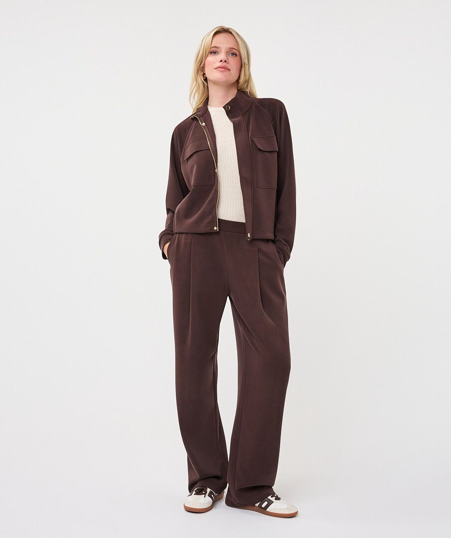 Full-length view of a woman wearing a chocolate brown utility jacket with matching wide-leg trousers, cream sneakers, and brown accents.