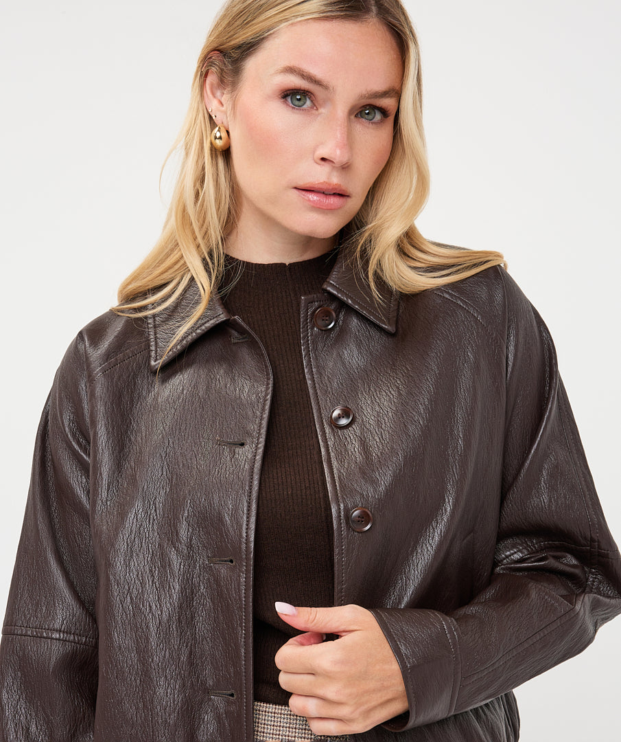Woman wearing a dark brown cropped faux leather jacket with a collared neckline and button-front closure, styled with a brown knit top