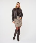 Woman wearing a dark brown cropped faux leather jacket with a collared neckline and button-front closure, styled with a brown knit top, patterned mini skirt, tights, and knee-high boots.