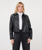 Woman wearing a dark gray cropped faux leather jacket with a collared neckline and button-front closure, layered over a white button-up shirt and paired with black straight-leg jeans 