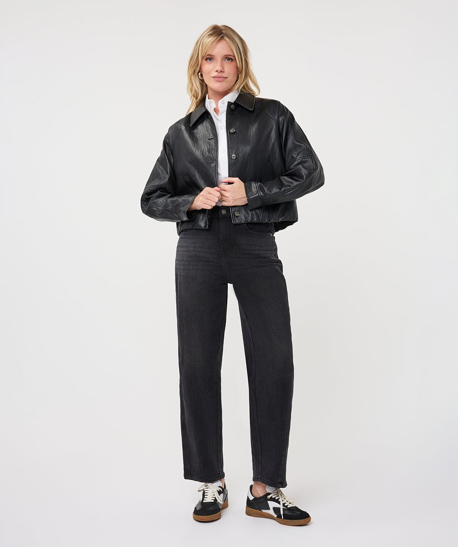  Woman wearing a dark gray cropped faux leather jacket with a collared neckline and button-front closure, layered over a white button-up shirt and paired with black straight-leg jeans and black-and-white sneakers.