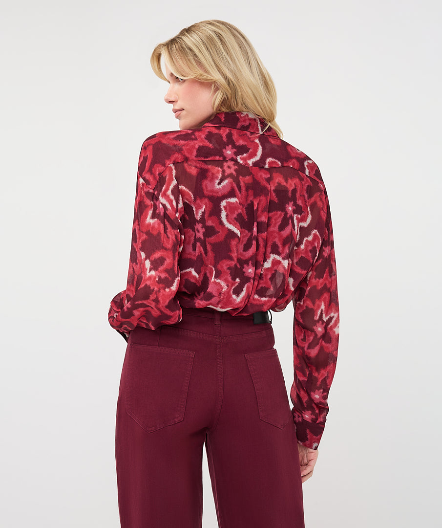 Back view of woman wearing a burgundy and red abstract print blouse with a relaxed fit and long sleeves, tucked into high-waisted burgundy pants.