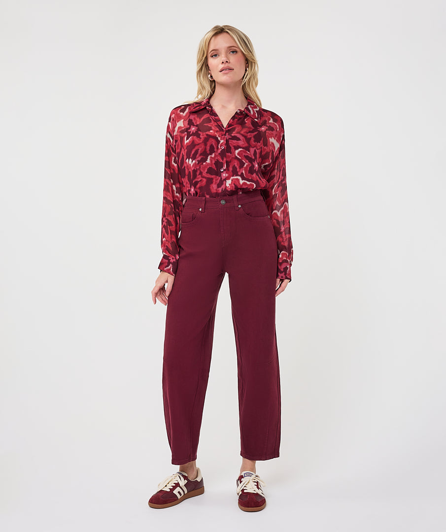 Woman wearing a burgundy and red abstract print blouse with long sleeves, paired with high-waisted burgundy straight-leg pants and burgundy-and-white sneakers.