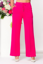 Woman wearing fuchsia wide-leg lounge pants with a drawstring waist, standing in a minimalist studio with floral arrangements behind her
