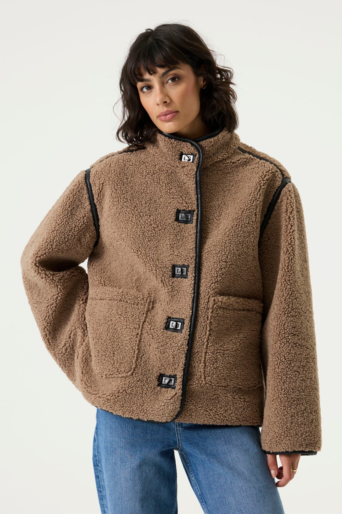 oman wearing a brown teddy fleece jacket with a high collar, toggle closures, contrast trim, and patch pockets, paired with blue jeans.