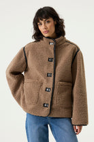 oman wearing a brown teddy fleece jacket with a high collar, toggle closures, contrast trim, and patch pockets, paired with blue jeans.