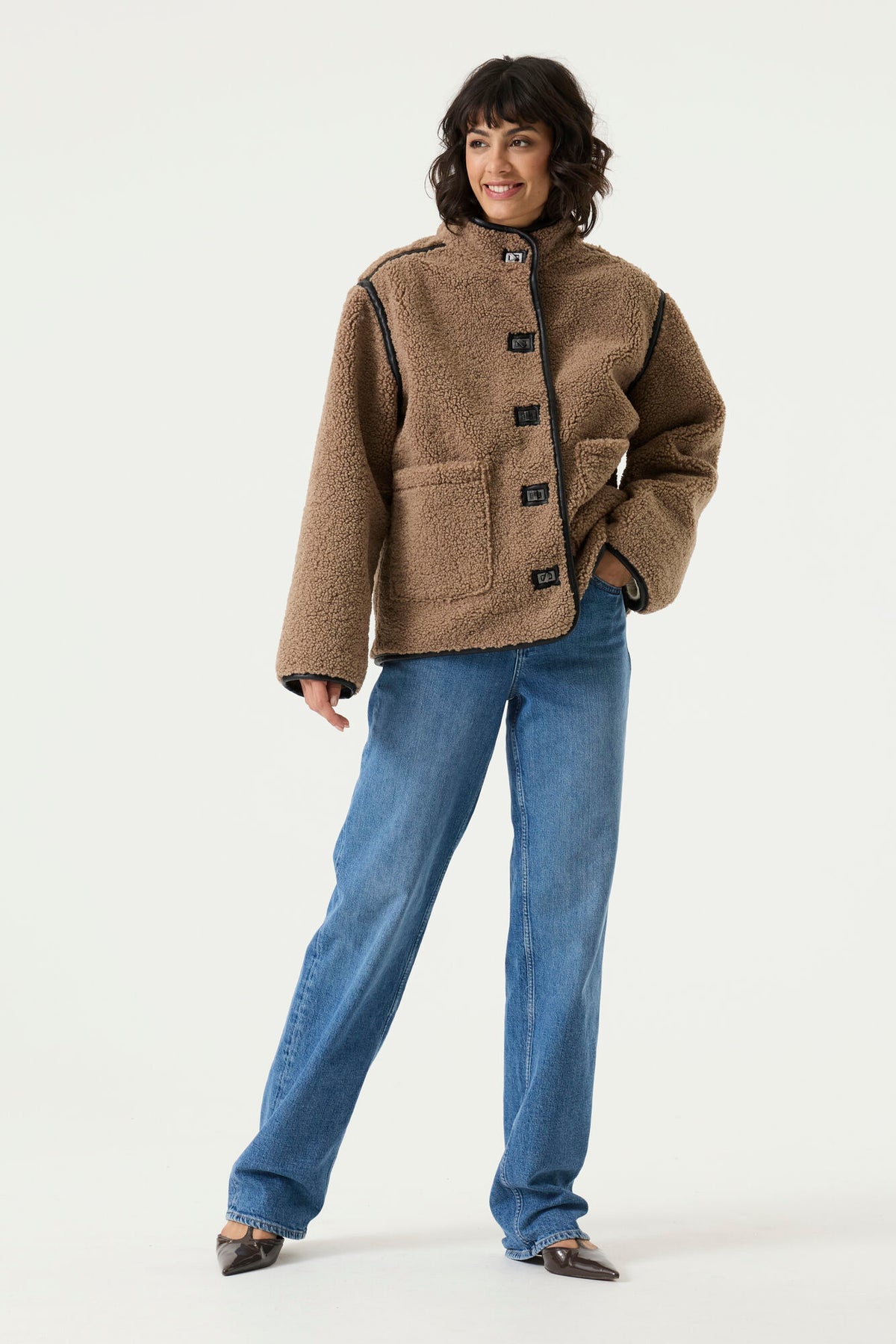 Woman wearing a brown teddy fleece jacket with a high collar, toggle closures, contrast trim, and patch pockets, paired with blue jeans.