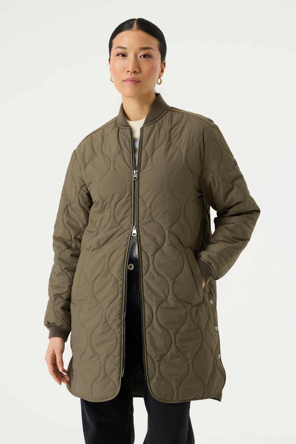 Woman modelling a longline olive green quilted jacket with a ribbed collar and cuffs, full front zip closure, side pockets, and rounded hem.