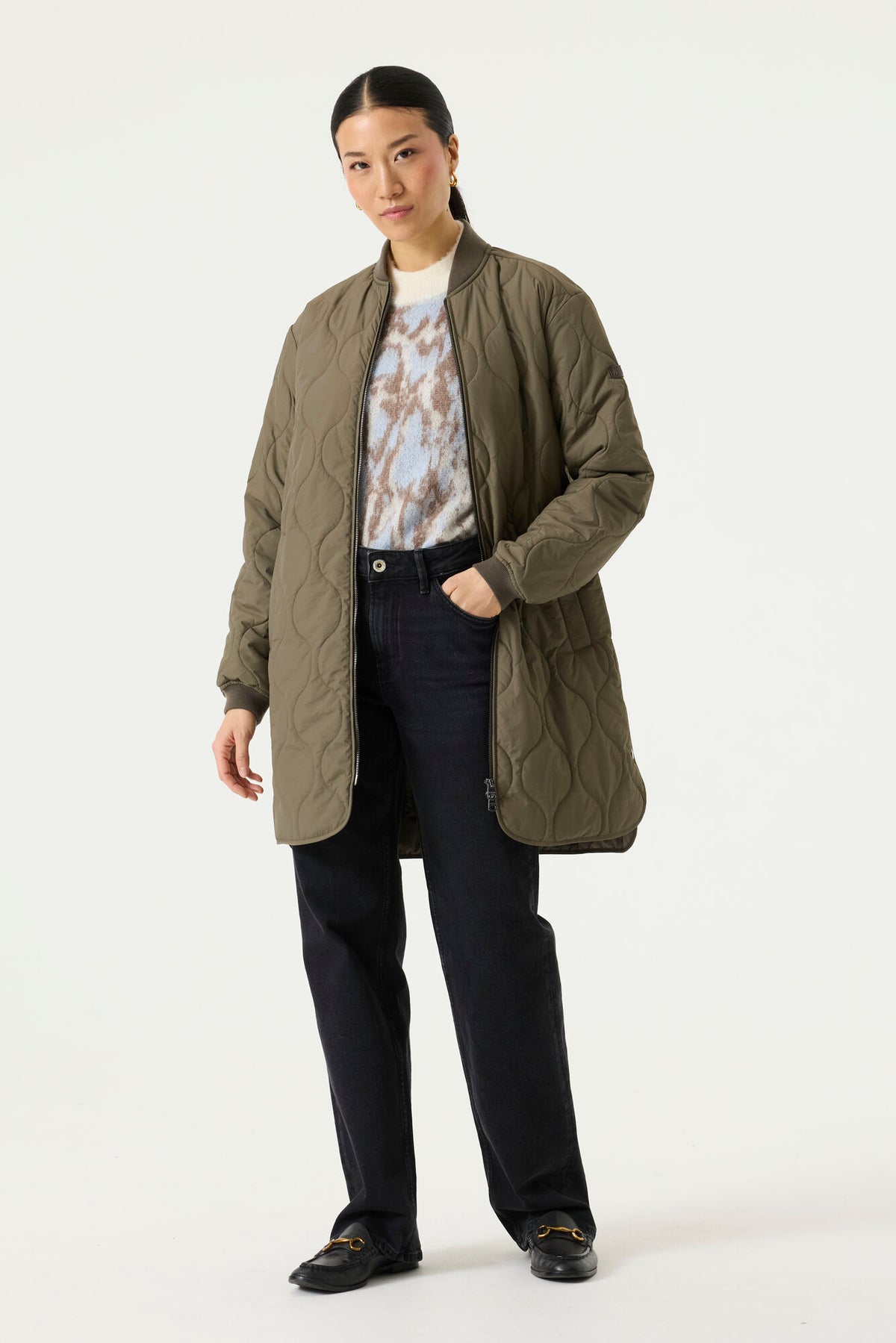 Woman modelling a longline olive green quilted jacket with a ribbed collar and cuffs, full front zip closure, side pockets, and rounded hem.