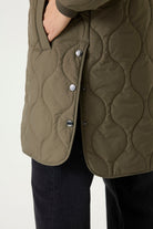 Woman modelling a longline olive green quilted jacket with a ribbed collar and cuffs, full front zip closure, side pockets, and rounded hem.