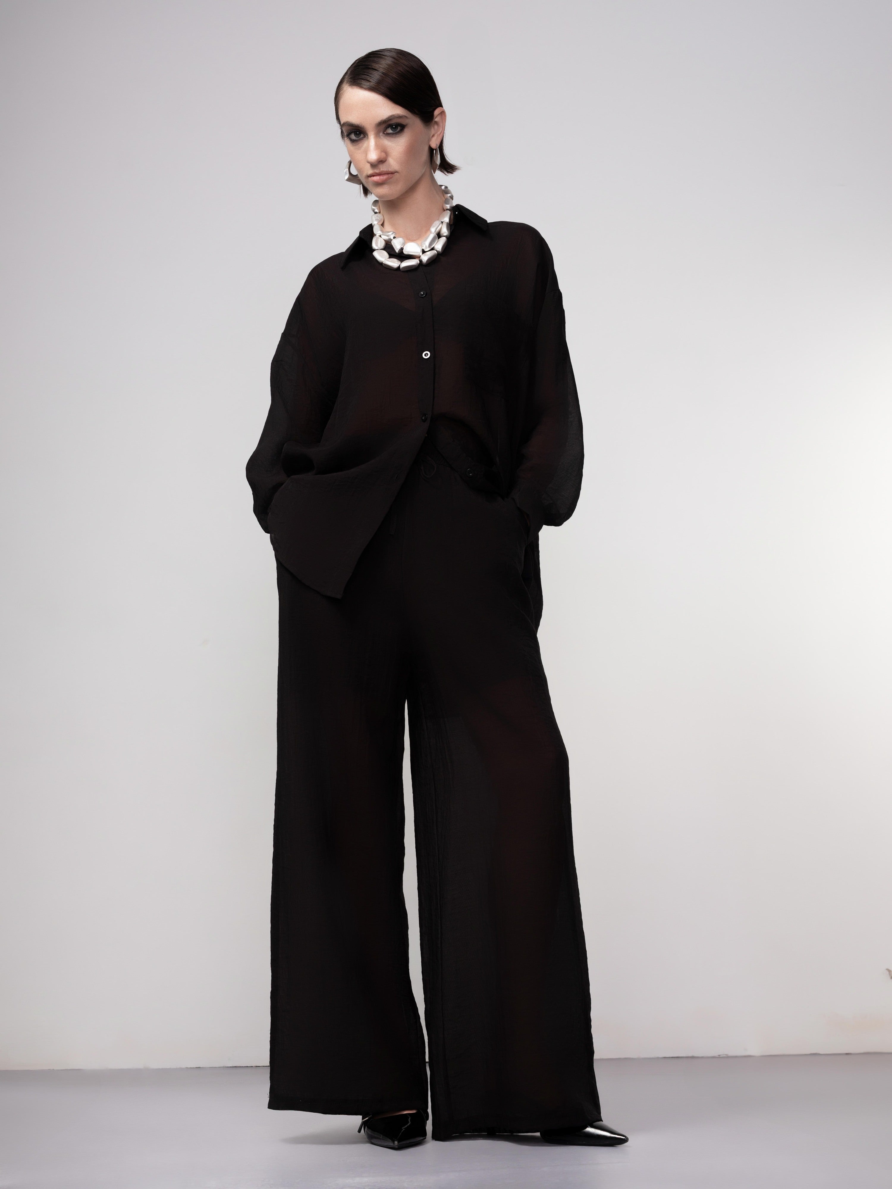 Woman wearing a black sheer button-front blouse with a relaxed fit, classic collar, and long sleeves, styled with wide-leg trousers and a statement necklace. 