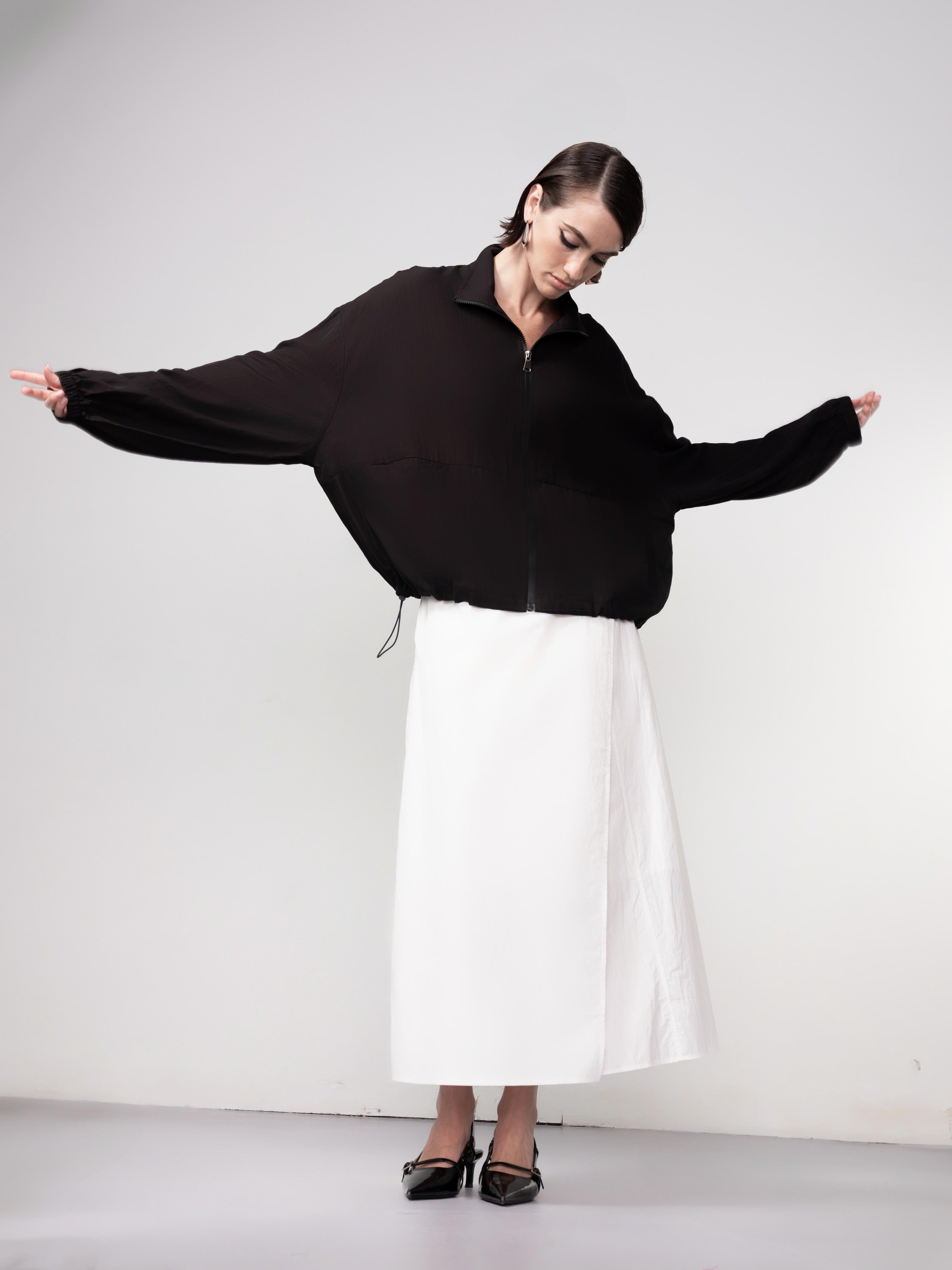 Model wearing a black relaxed-fit zip blouse with voluminous sleeves and a white midi skirt, posing with arms extended.