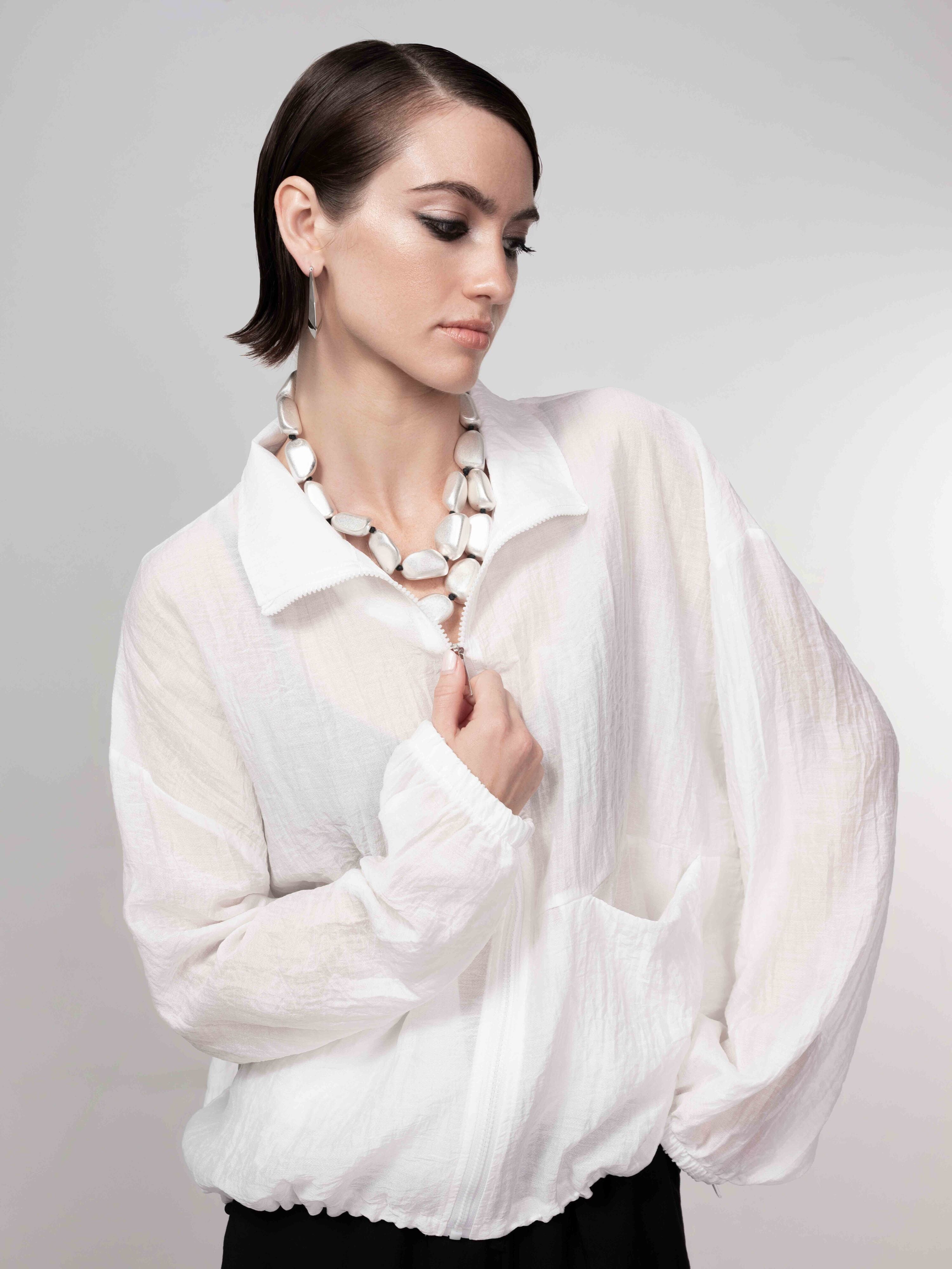 Close-up of model wearing a white sheer zip-front blouse with long sleeves, styled with a statement necklace and black bottoms.