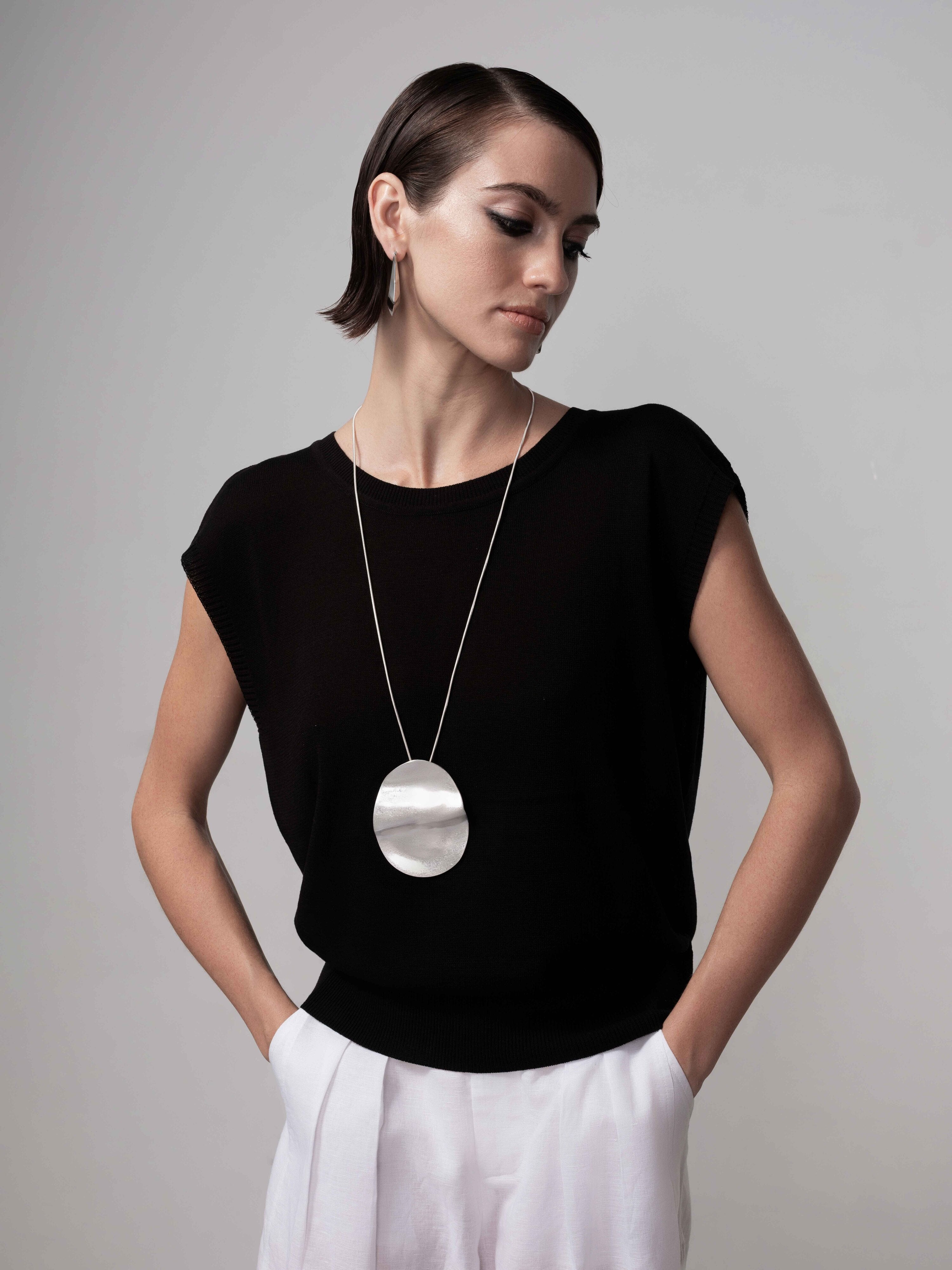 Model wearing a black cap sleeve knit sweater with a relaxed silhouette, paired with light trousers and a long pendant necklace, hands in pockets.