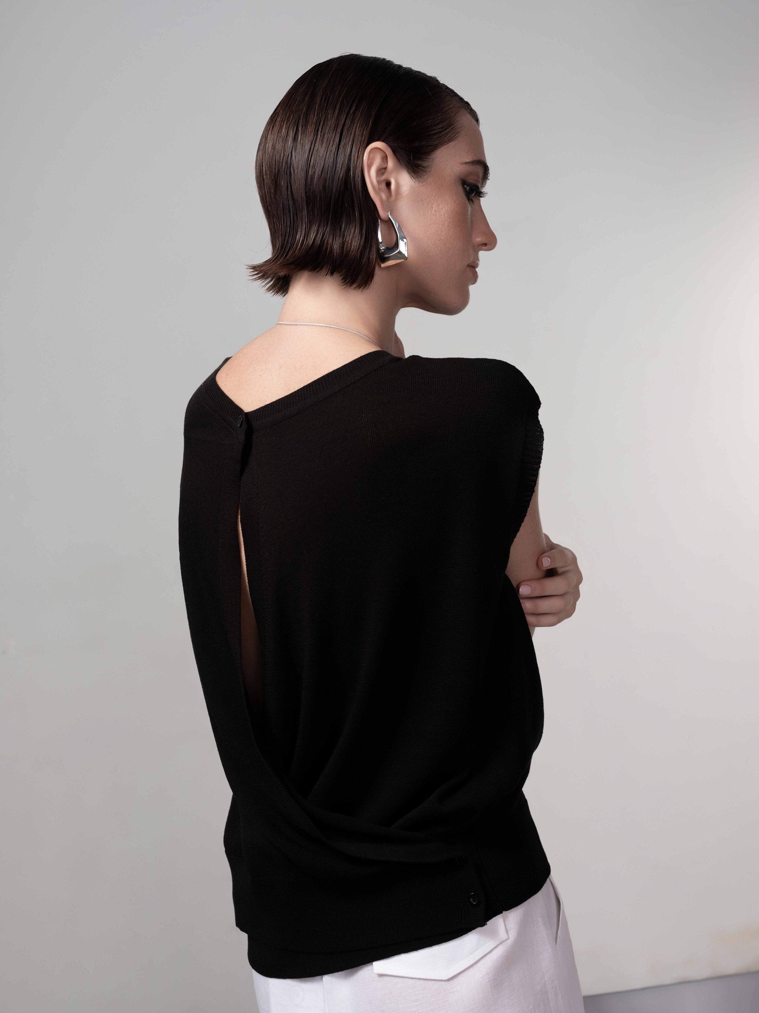 Back view of a black cap sleeve knit sweater featuring a relaxed drape and subtle back slit detail, styled with light trousers.