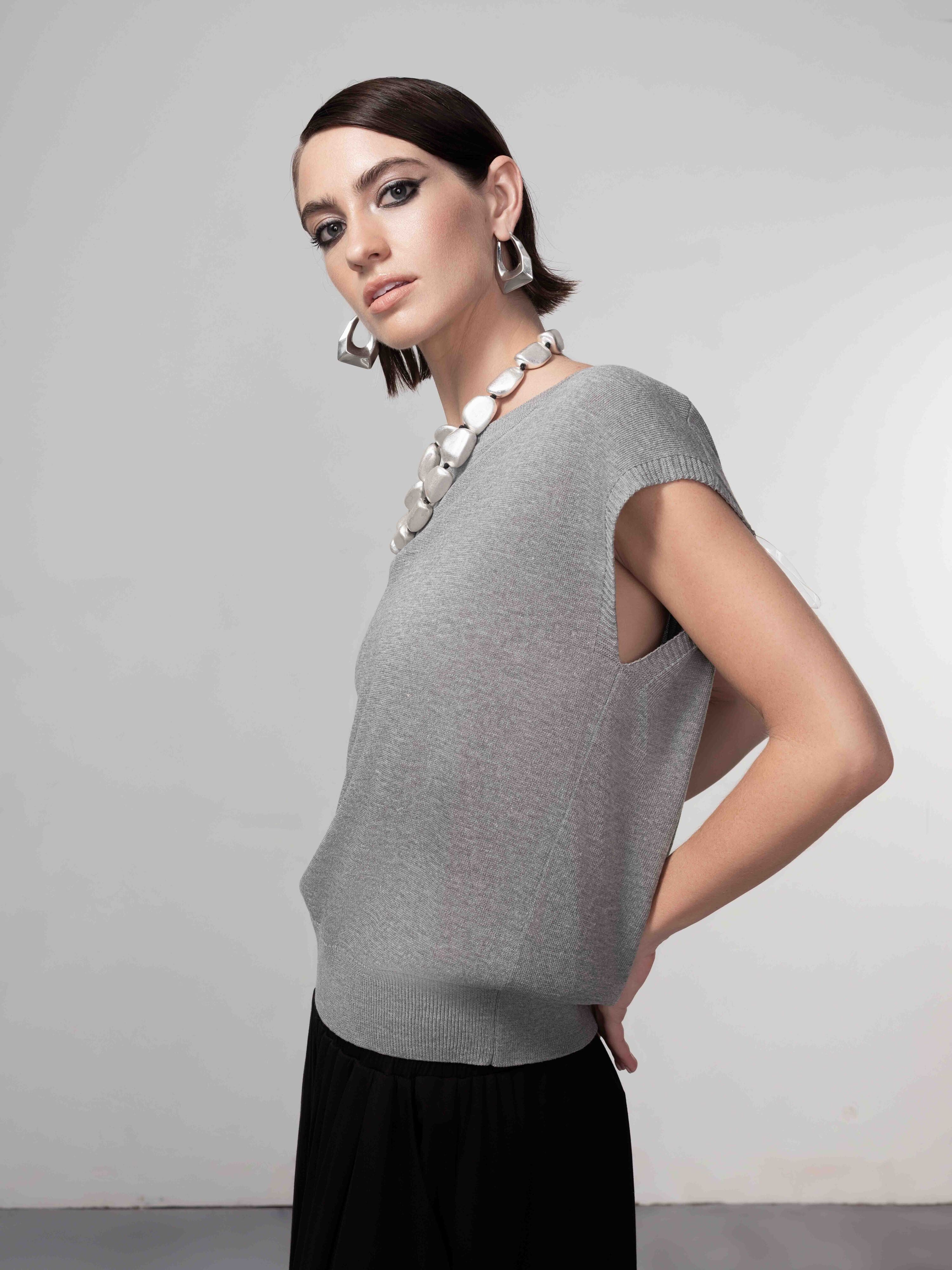 Model wearing a grey cap sleeve knit sweater with a relaxed draped fit, styled with a statement necklace and dark skirt, shown in side profile.