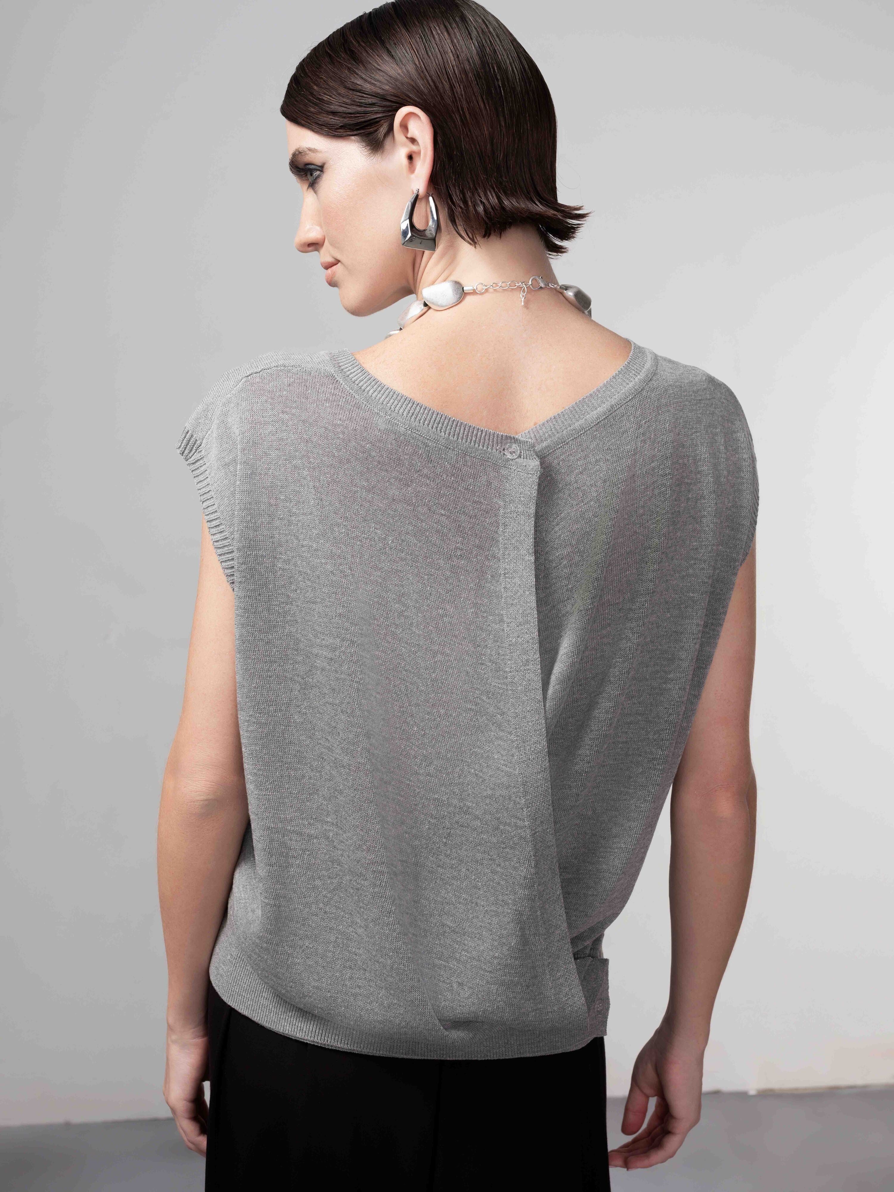 Model wearing a grey cap sleeve knit sweater with a relaxed draped fit, styled with a statement necklace and dark skirt