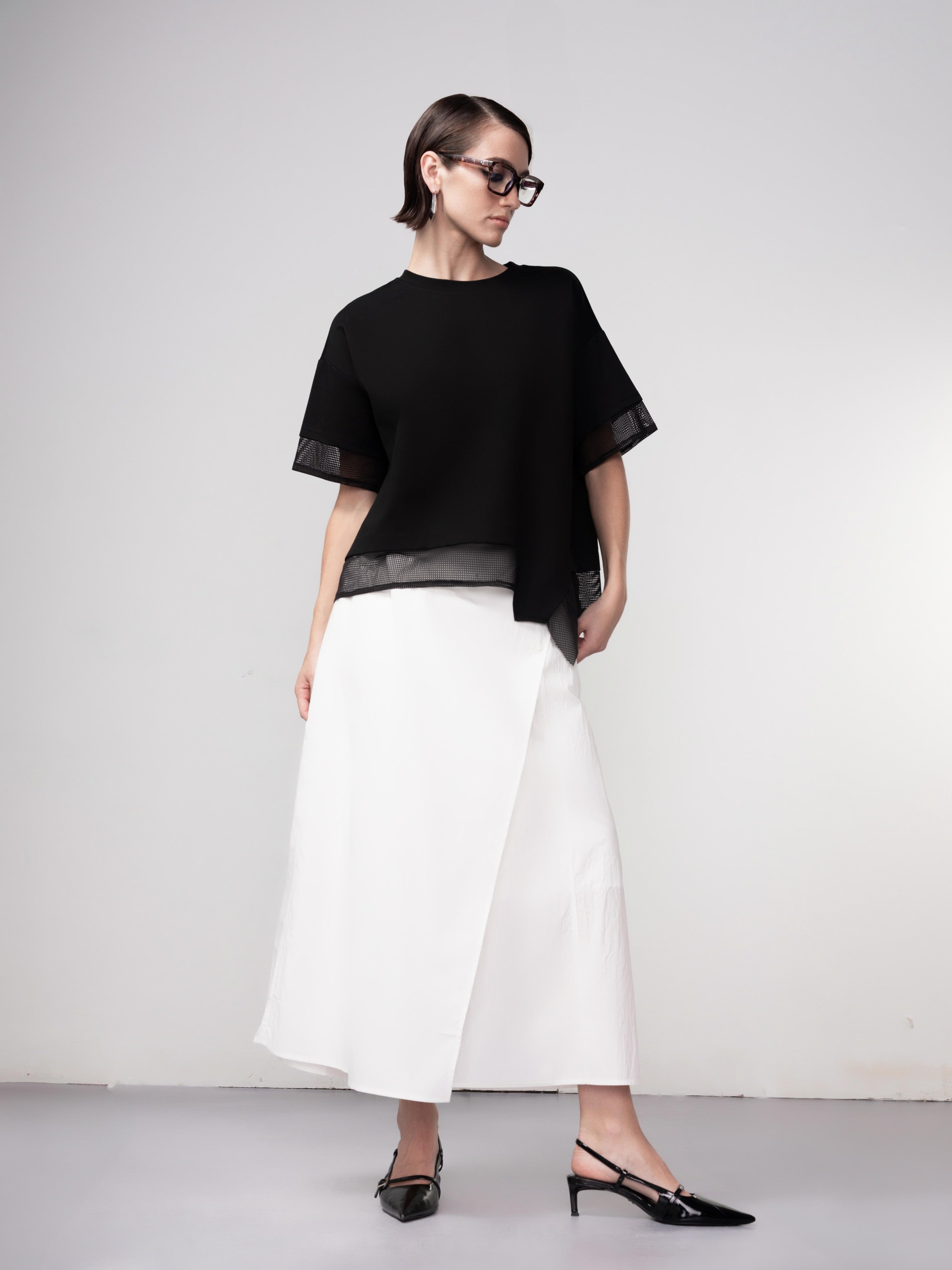 Full-length view of a model wearing a black relaxed-fit blouse with sheer mesh trim at the sleeves and hem, paired with a white midi skirt and pointed slingback heels.