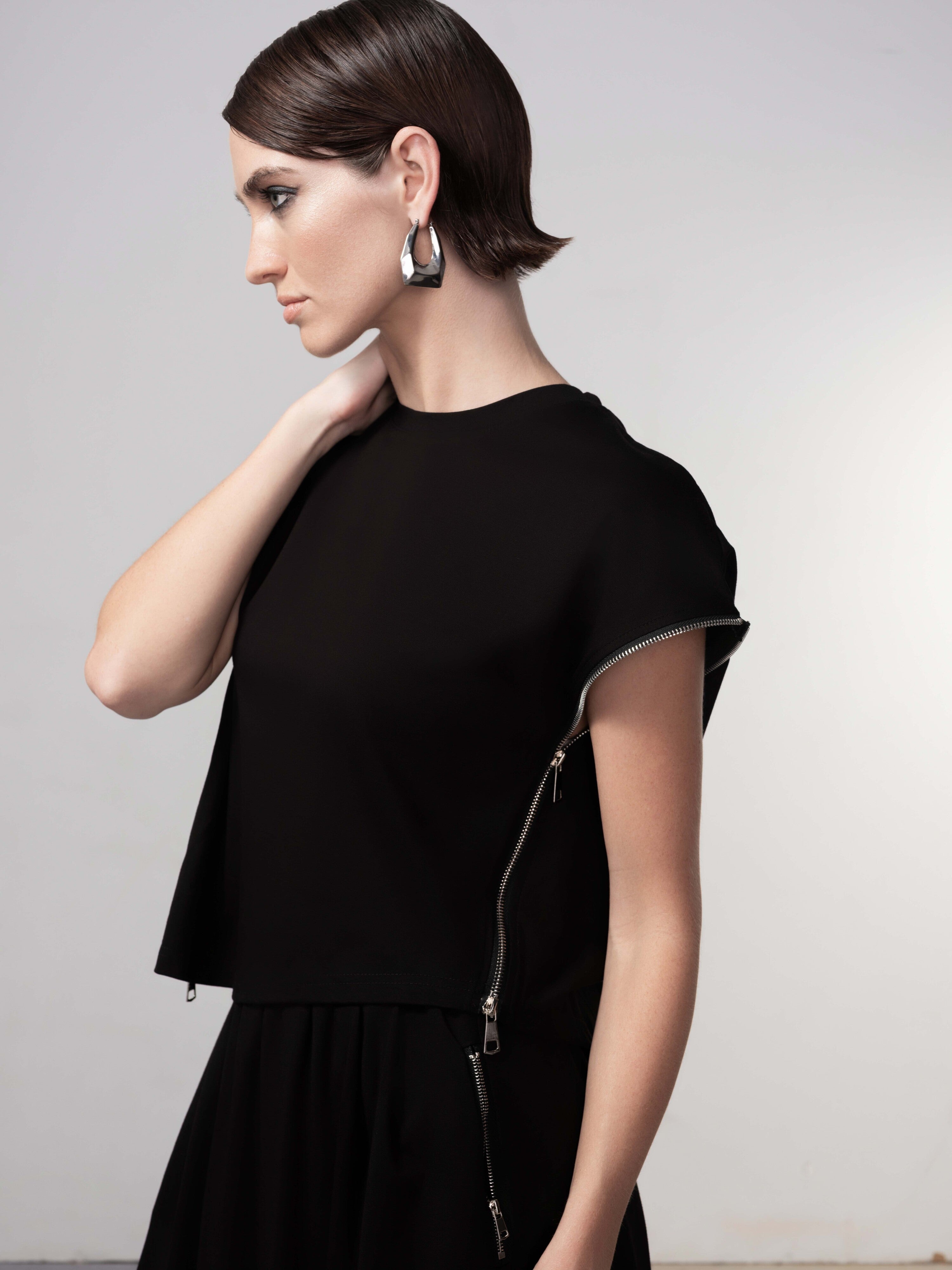Side profile view of a model wearing a black cropped pullover with cap sleeves and silver zip detailing along the side, styled with statement earrings.