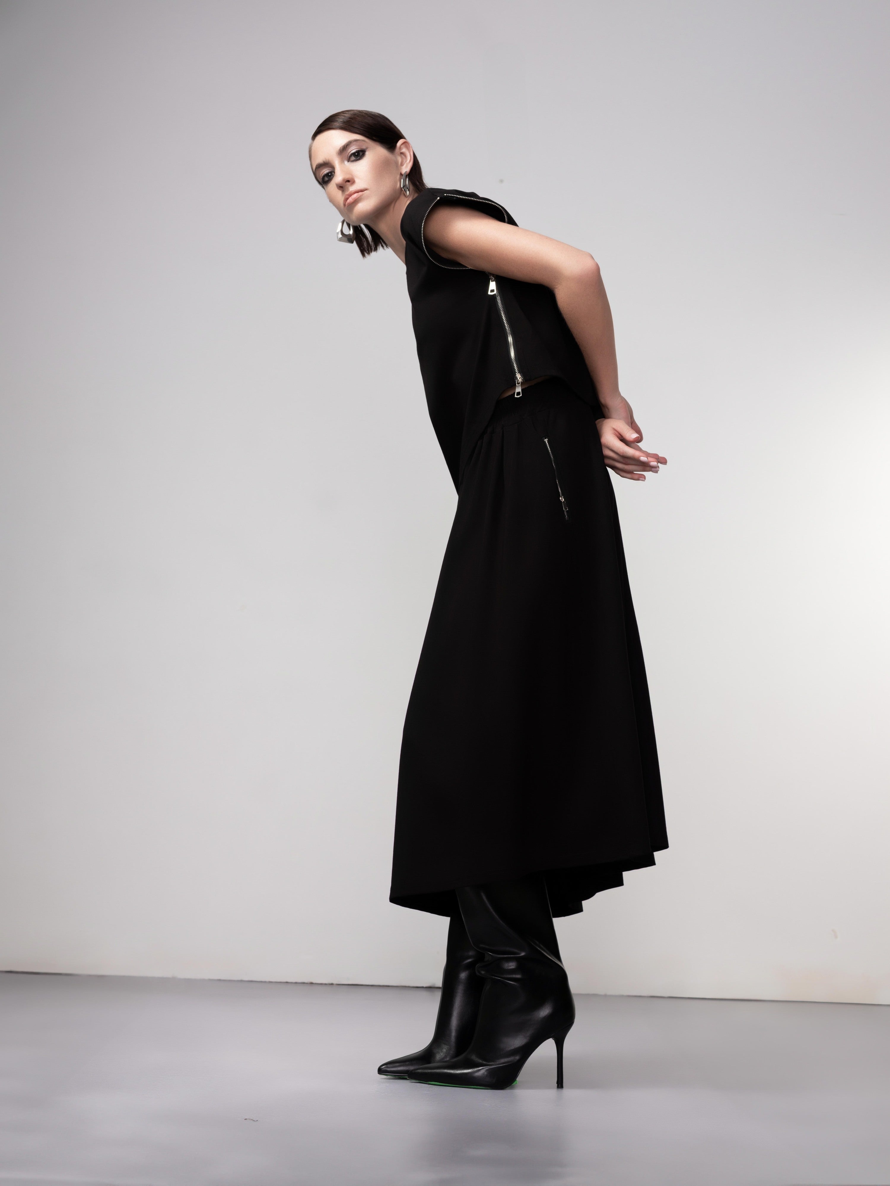 Full-length view of a model wearing a black cropped pullover with side zip accents, paired with a matching midi skirt and pointed high-heeled boots, posed in a modern studio setting.