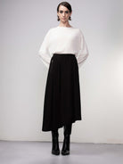 A woman wearing a white long-sleeve top paired with a black asymmetrical midi skirt. She is styled with knee-high black boots and minimal accessories. The skirt has a smooth, flowing fabric that drapes elegantly, featuring an elastic waistband for a comfortable fit.