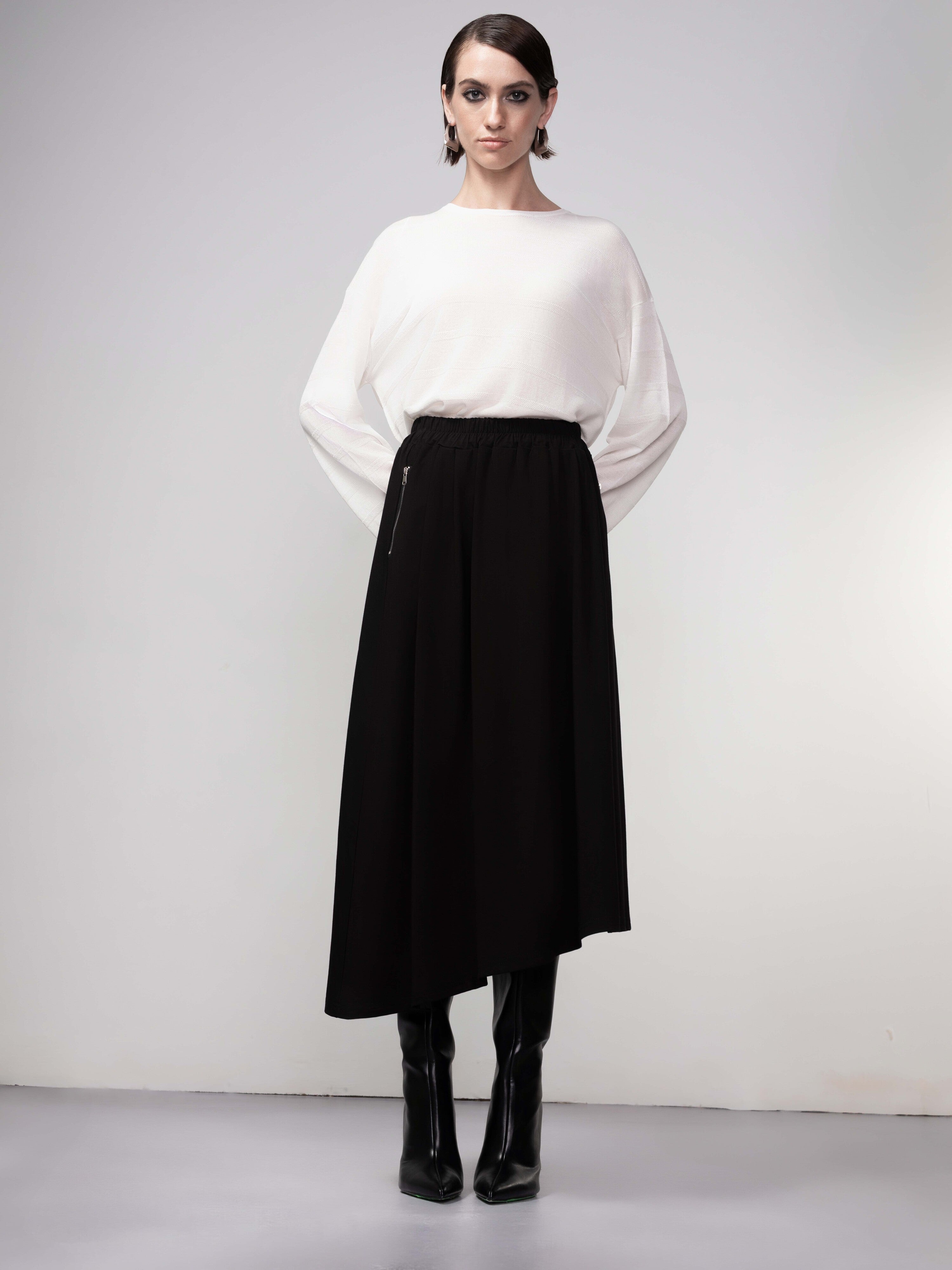 A woman wearing a white long-sleeve top paired with a black asymmetrical midi skirt. She is styled with knee-high black boots and minimal accessories. The skirt has a smooth, flowing fabric that drapes elegantly, featuring an elastic waistband for a comfortable fit.